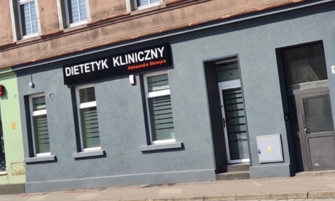 Fasada budynku z szyldem 'Dietetyk Kliniczny Aleksandra Skowyra', okna z żaluzjami i dwoje drzwi wejściowych.