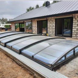 STELLER PPHU Tomasz Steller - Montaż zadaszenia basenu z szarej konstrukcji na betonowej podstawie, widoczny częściowo wypełniony basen i budynek w tle w trakcie budowy.