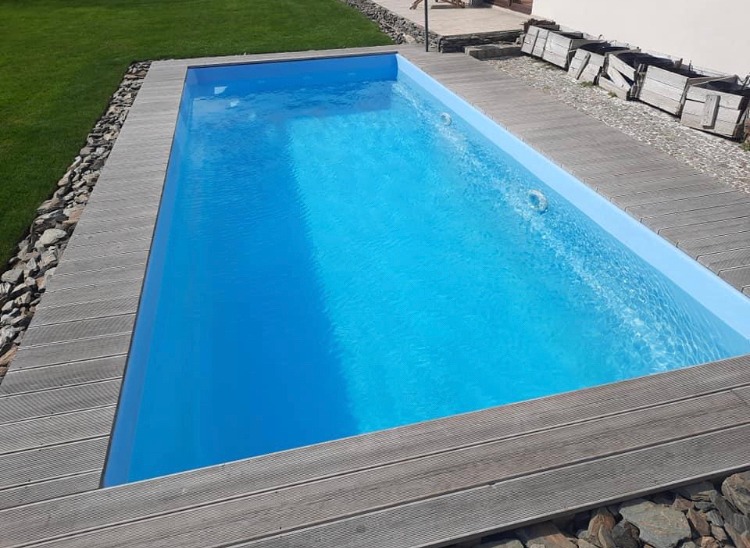 Prostokątny, gotowy basen ogrodowy z drewnianym obramowaniem i kamieniami dekoracyjnymi, wypełniony błękitną wodą, widoczne dysze do hydromasażu.