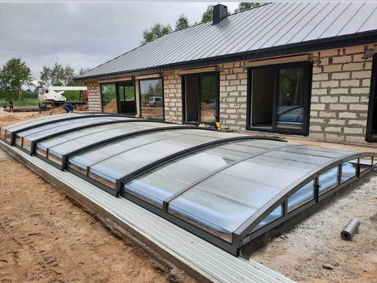 Montaż zadaszenia basenu z szarej konstrukcji na betonowej podstawie, widoczny częściowo wypełniony basen i budynek w tle w trakcie budowy.