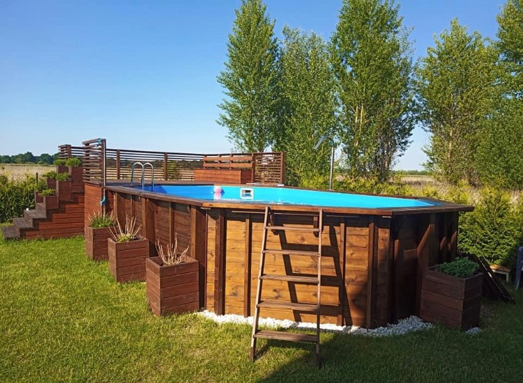 Drewniany basen naziemny z drabinką, schodami i prysznicem, otoczony kostką granitową i donicami z roślinami, na tle zielonego trawnika i drzew