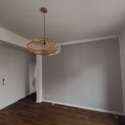 Pracownia projektowo-remontowa „Kreator” Mateusz Langosz - Szara ściana w świeżo wykończonym pokoju z drewnianą podłogą, ozdobnymi listwami przysufitowymi i przypodłogowymi oraz wiszącą lampą z wikliny