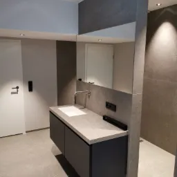 Nowoczesna łazienka z szarą armaturą, umywalką wpuszczaną w blat i dużym lustrem, wykończona płytkami w odcieniach szarości.