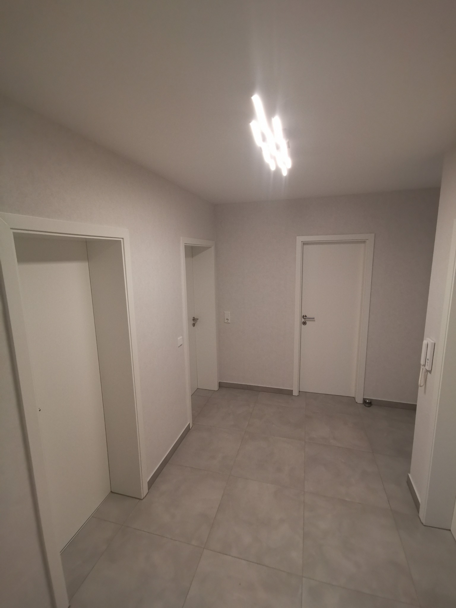 Wnętrze korytarza z trzema białymi drzwiami, szarą podłogą z płytek i nowoczesnym oświetleniem sufitowym.