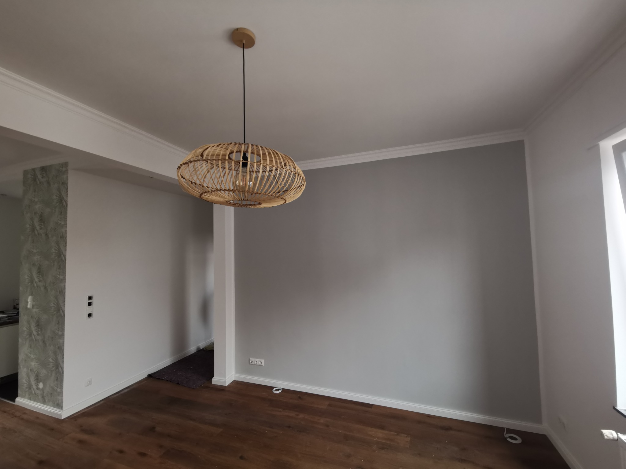 Szara ściana w świeżo wykończonym pokoju z drewnianą podłogą, ozdobnymi listwami przysufitowymi i przypodłogowymi oraz wiszącą lampą z wikliny