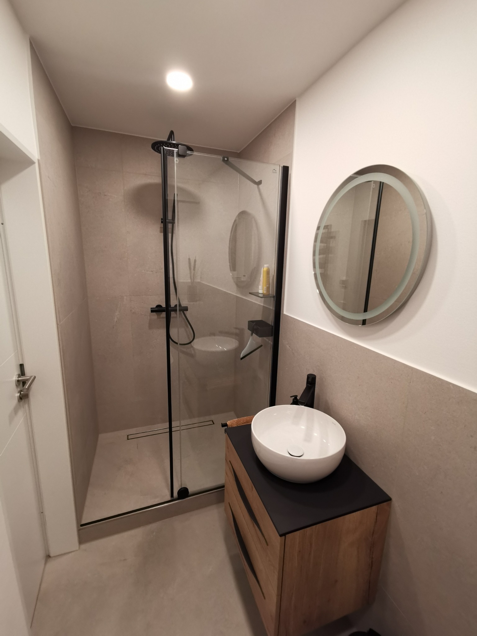 Nowoczesna łazienka z kabiną prysznicową typu walk-in z czarnymi ramami, okrągłym lustrem z podświetleniem LED, umywalką nablatową na drewnianej szafce z czarnym blatem i szarą glazurą.