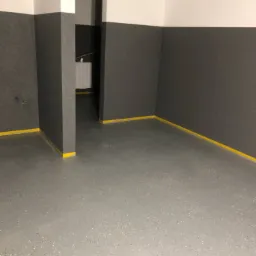 Pokój po malowaniu ścian na szaro z białym pasem u góry, zabezpieczony żółtą taśmą malarską przy podłodze i ościeżnicy, podłoga w jasnym odcieniu szarości z ciemnymi drobinkami.