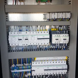 Rozdzielnica elektryczna z widocznymi bezpiecznikami, stycznikami i przewodami oznaczonymi numerami, zamontowana w metalowej obudowie.