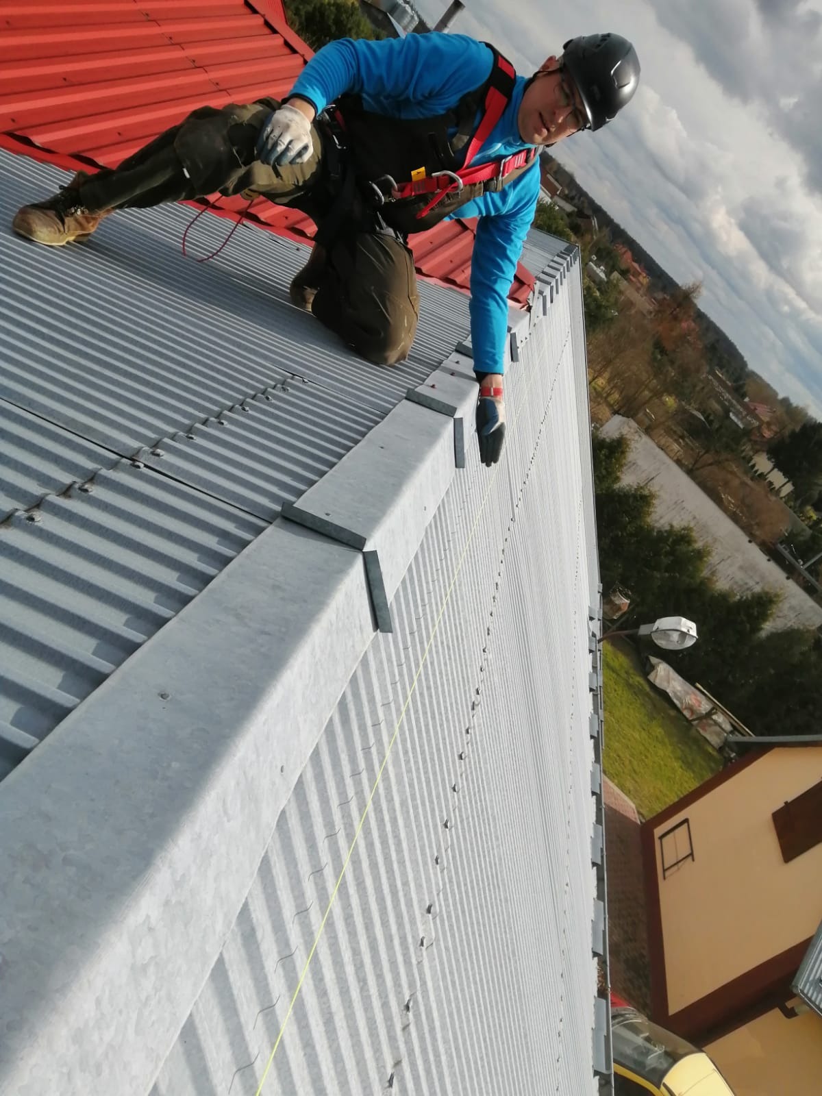 Pracownik w uprzęży zabezpieczającej montuje elementy na metalowym dachu z wyraźnymi żłobieniami, widoczne narzędzia i lina asekuracyjna, w tle zabudowania i pochmurne niebo.