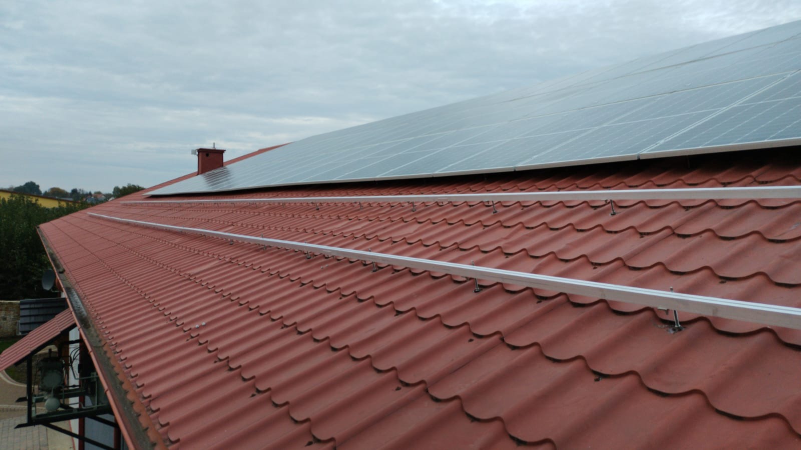 Panele słoneczne zamontowane na czerwonym dachu z dachówki ceramicznej, widoczne szyny montażowe i mocowania, pochmurne niebo w tle.