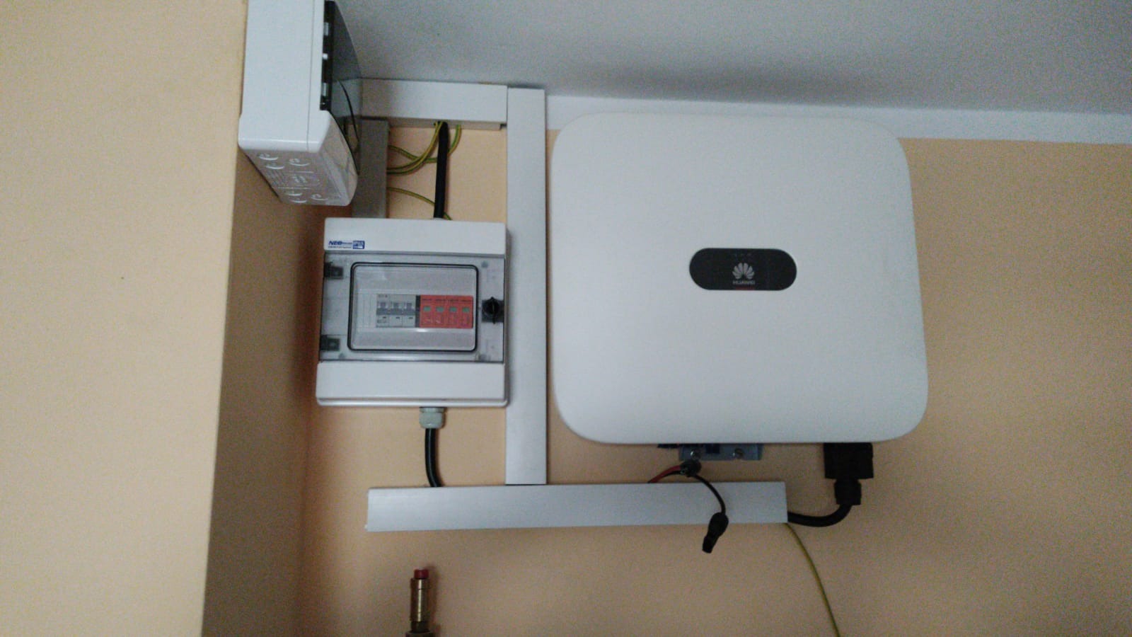 Biały inwerter Huawei zamontowany na ścianie obok skrzynki bezpiecznikowej z widocznymi przewodami i elementami montażowymi.