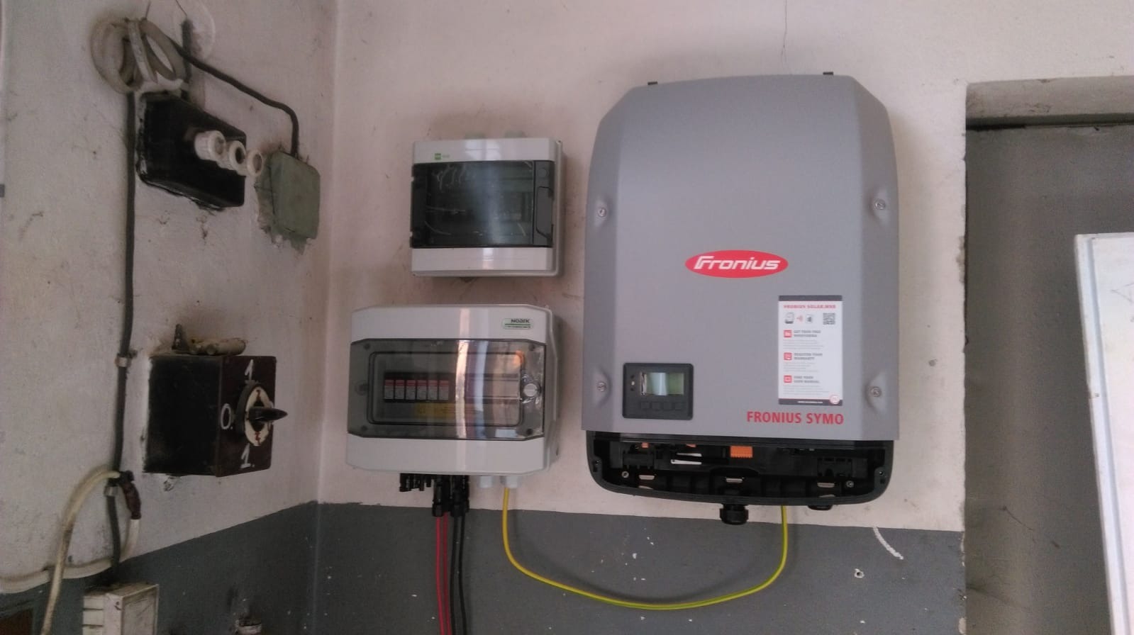 Szary inwerter Fronius Symo zamontowany na szarej, pomalowanej do połowy ścianie w pomieszczeniu z widoczną starą instalacją elektryczną i skrzynkami rozdzielczymi.