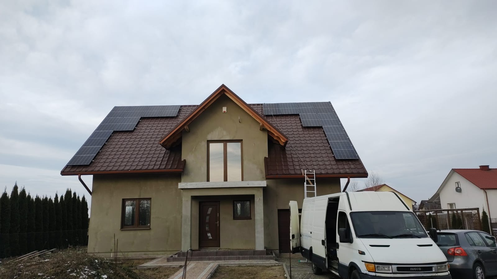 Dom jednorodzinny z brązowym dachem, na którym zamontowane są panele fotowoltaiczne; obok zaparkowany biały samochód dostawczy z otwartymi drzwiami.