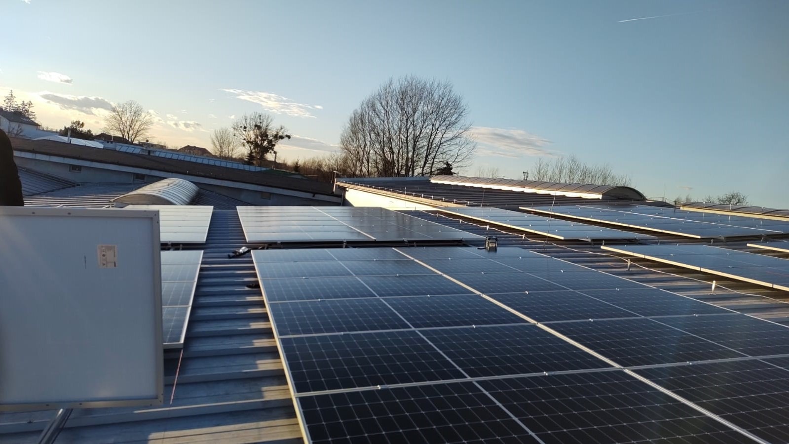 Montaż paneli słonecznych na dachu hali przemysłowej, widoczny fragment instalacji z jednym panelem odwróconym tyłem, błękitne niebo w tle.