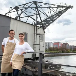 Uśmiechnięta para w beżowych fartuchach roboczych na tle starego dźwigu portowego i panoramy miasta z rzeką.