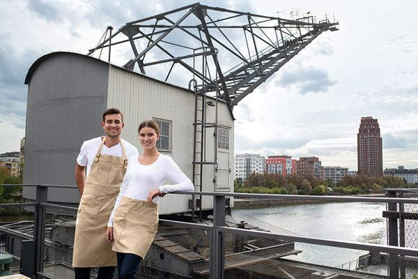 Uśmiechnięta para w beżowych fartuchach roboczych na tle starego dźwigu portowego i panoramy miasta z rzeką.