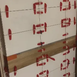 Świeżo ułożone, białe płytki z czerwonymi klinami dystansowymi, częściowo przysłonięte drewnopodobną płytką w trakcie prac wykończeniowych.