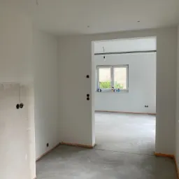 Wnętrze w trakcie przygotowania do prac wykończeniowych: ściany częściowo zagruntowane, widoczne puszki elektryczne, surowa wylewka podłogowa, listwy przypodłogowe w narożnikach, okno z białym...