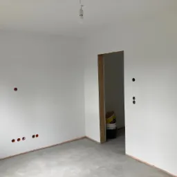 Wnętrze w trakcie prac wykończeniowych: świeżo pomalowane białe ściany z widocznymi puszkami instalacyjnymi, surowy beton na podłodze zabezpieczony taśmą, widoczna ościeżnica drzwi i wiadro z farbą...