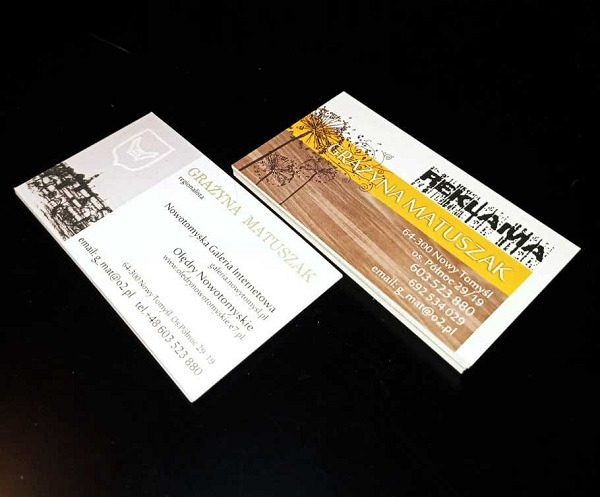 Dwie wizytówki firmy Grażyna Matuszak: jedna z grafiką Nowego Tomyśla i herbem, druga z motywem drewna i kwiatów, na czarnym tle.