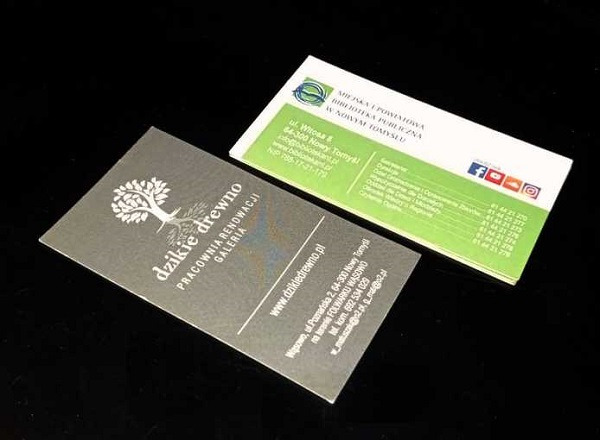 Dwie wizytówki leżące na czarnym tle: jedna z logo firmy 'Dzikie drewno' z motywem drzewa i adresem, druga z logo Miejskiej i Powiatowej Biblioteki Publicznej w Nowym Tomyślu, z zielonym paskiem...