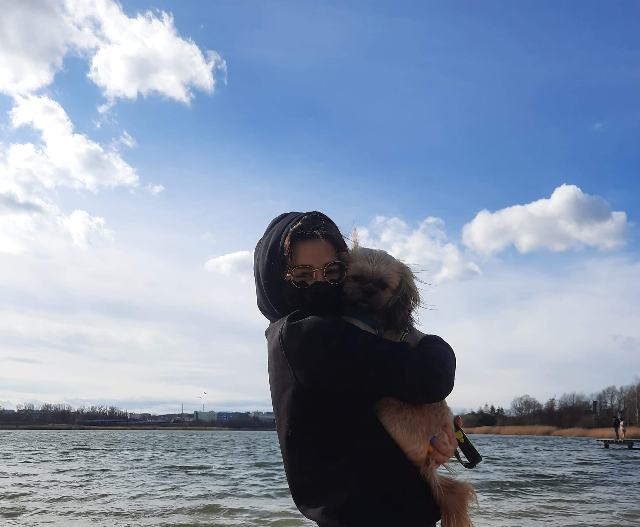 Osoba w czarnej bluzie z kapturem i okularach trzyma na rękach małego, jasnego psa z niebieską obrożą na tle jeziora i błękitnego nieba z chmurami.