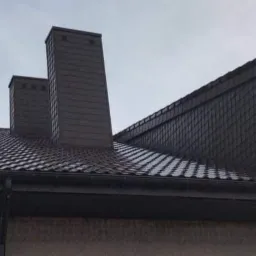 Dom z nowym, brązowym dachem z blachodachówki, dwoma kominami obłożonymi panelami oraz zamontowanymi rynnami w kolorze dachu, na tle pochmurnego nieba.