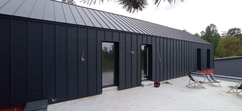 Nowoczesny dom z ciemną, metalową elewacją i dachem panelowym, widoczne niedokończone obróbki blacharskie i wystające kable, na dachu leżaki.