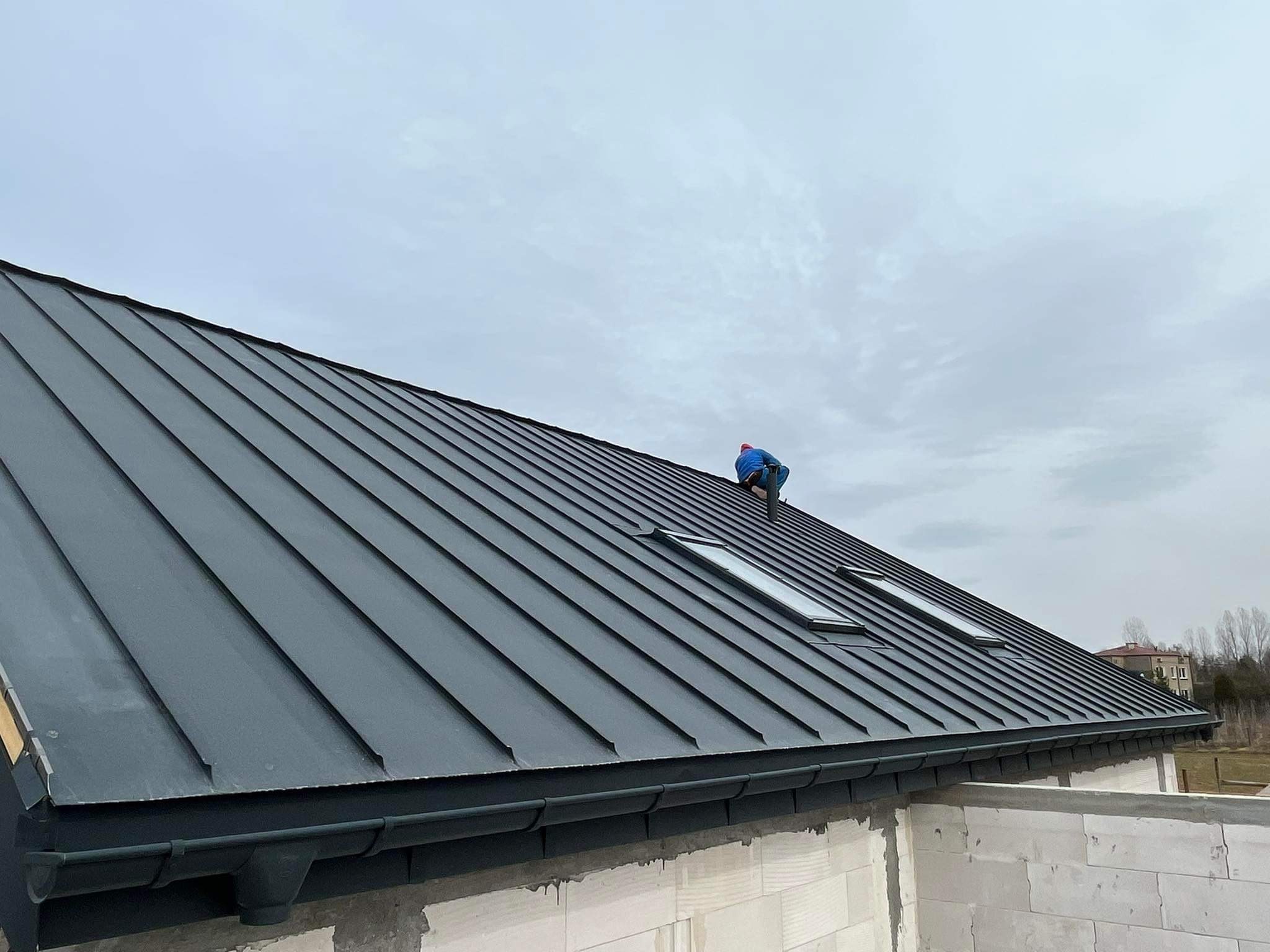 Szary dach z blachy na ukończonym budynku, widoczne okna dachowe i system rynnowy, dekarz w niebieskim ubraniu w trakcie pracy na dachu.