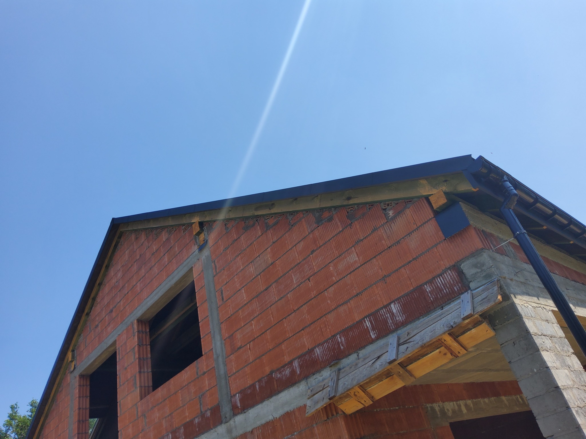 Budowa domu z czerwonej cegły ceramicznej, widoczne obróbki blacharskie i rynna w kolorze grafitowym na tle błękitnego nieba.