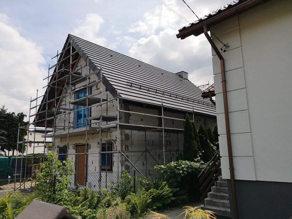 Dom w trakcie budowy z nowym dachem z czarnej dachówki, otoczony rusztowaniem, widoczne okna zabezpieczone folią.