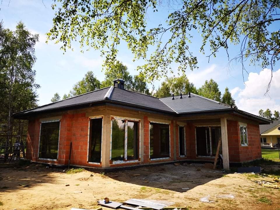Dom jednorodzinny w trakcie budowy z nowym, ciemnoszarym dachem z dachówki ceramicznej, widok z zewnątrz na surowy stan budynku z niezabudowanymi oknami i elementami obróbki blacharskiej.