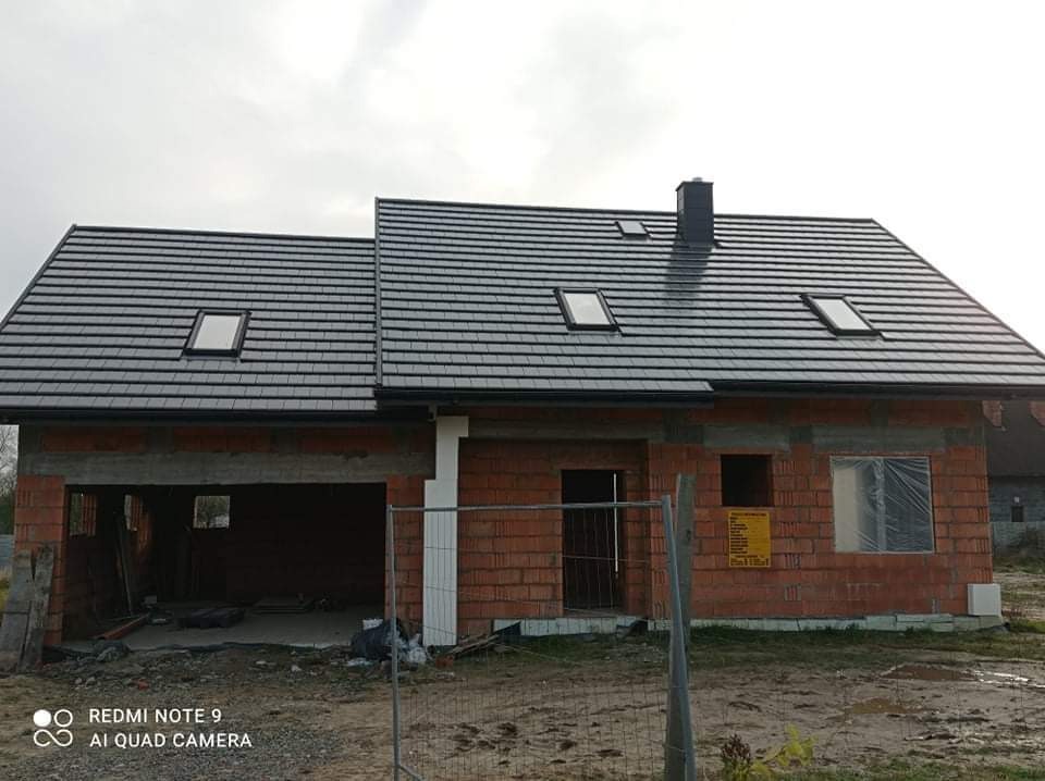 Dom w trakcie budowy z nowym, ciemnoszarym dachem z dachówki ceramicznej i oknami dachowymi, widoczne obróbki blacharskie i system rynnowy, ściany z czerwonej cegły.