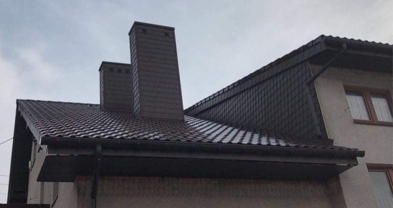 Dom z nowym, brązowym dachem z blachodachówki, dwoma kominami obłożonymi panelami oraz zamontowanymi rynnami w kolorze dachu, na tle pochmurnego nieba.