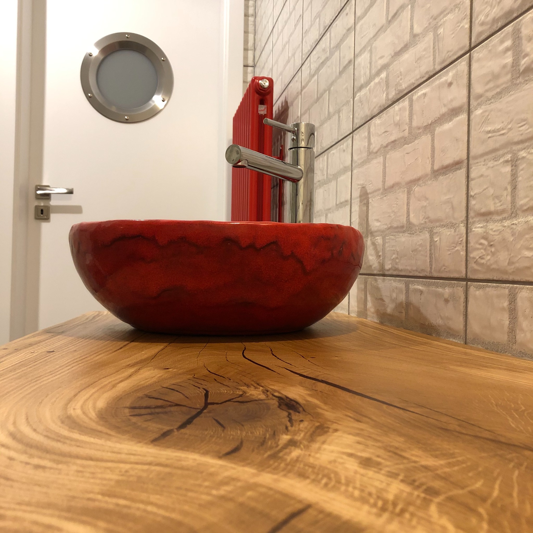 Czerwona, ceramiczna umywalka nablatowa na blacie z litego drewna o widocznych słojach, z baterią w tle, na ścianie biała cegła i czerwony grzejnik, w tle drzwi z okrągłym okienkiem.