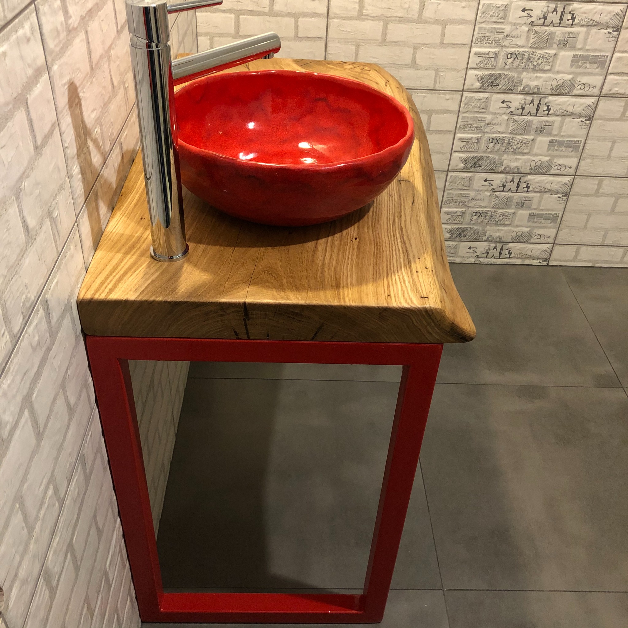 Czerwona, ceramiczna umywalka nablatowa z chromowaną baterią, osadzona na blacie z litego drewna z surowym brzegiem, wspartym na czerwonym, metalowym stelażu.