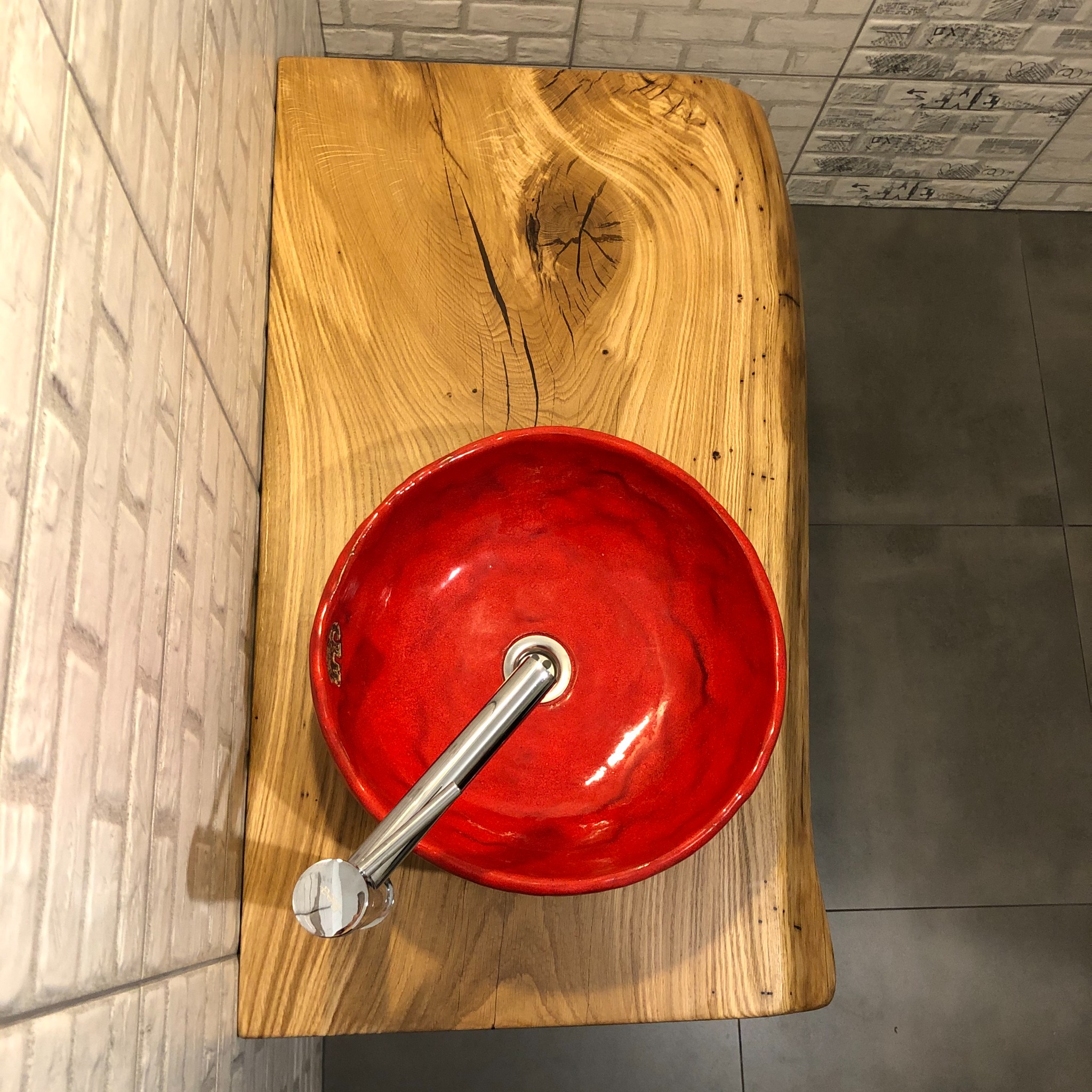 Czerwona, ceramiczna umywalka nablatowa z chromowaną baterią, osadzona na blacie z litego drewna dębowego z wyraźnym usłojeniem i sękami, na tle ściany wyłożonej białą cegłą i szarych płytek...