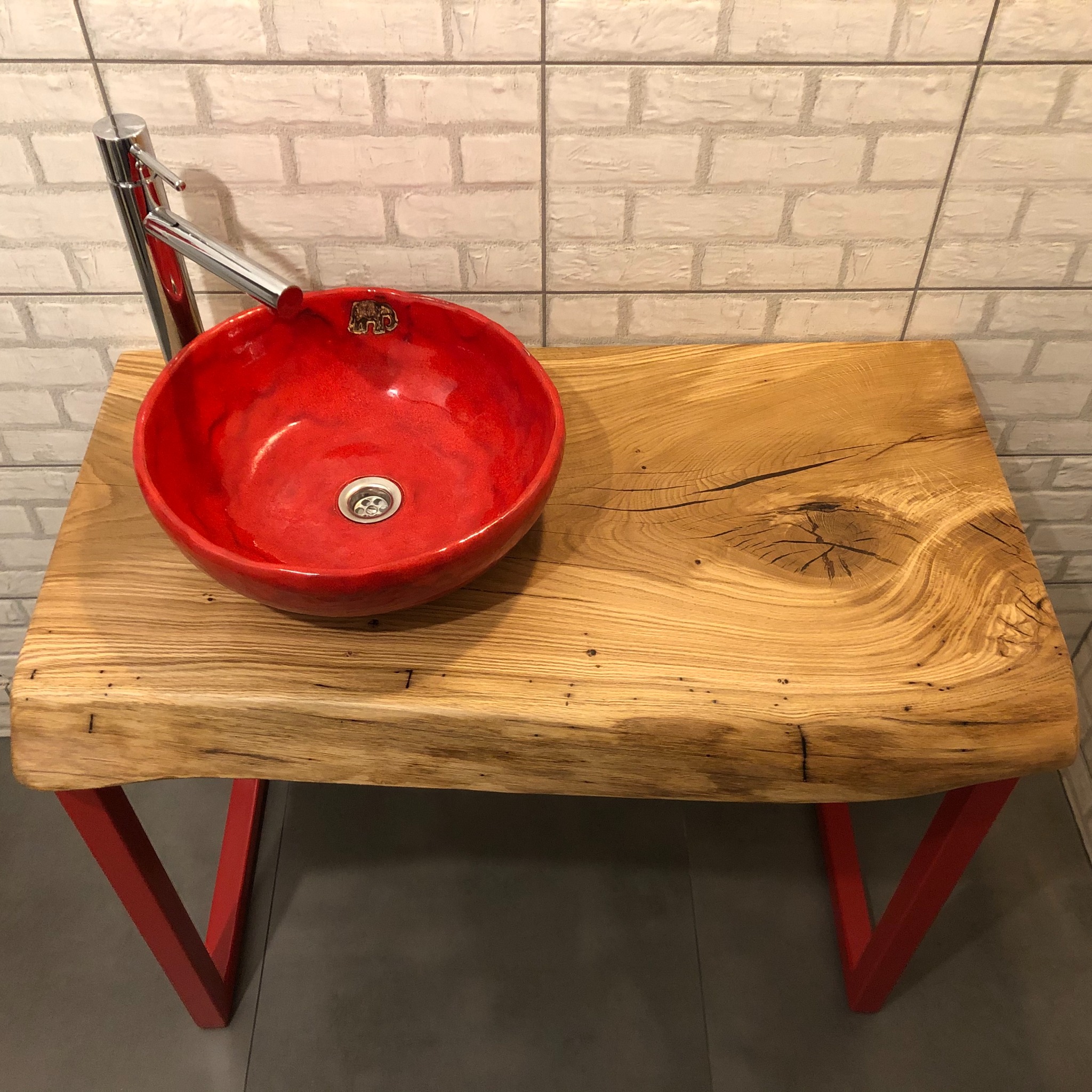 Umywalka ceramiczna w intensywnym czerwonym kolorze, osadzona na blacie z naturalnego drewna o wyraźnym usłojeniu i krawędzi typu live edge, wsparta na czerwonych, metalowych nogach w kształcie...