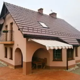 Dom jednorodzinny z beżową elewacją i brązowym dachem, z markizą w biało-brązowe paski nad wejściem do podcienia, gdzie stoi drewniany stół i krzesła.