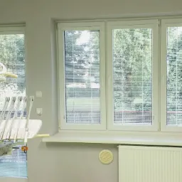 Żaluzje aluminiowe w oknie gabinetu dentystycznego w Tarnobrzegu, widoczne zieleń drzew za oknem, sprzęt dentystyczny po lewej stronie.