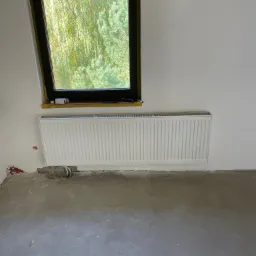 Biały, żebrowy grzejnik zamontowany pod oknem w surowym wnętrzu z betonową podłogą; widoczne rury doprowadzające instalację, fragment okna z widokiem na drzewa.