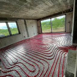 Czerwone rury ogrzewania podłogowego ułożone na folii w surowym wnętrzu z widokiem na zieleń za oknem, widoczne białe klipsy mocujące rury do siatki.