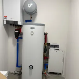 Biały kondensacyjny piec gazowy Viessmann wiszący na ścianie, obok pionowy biały zasobnik Nibe z szarym zbiornikiem wyrównawczym, połączone niebieskimi i czerwonymi rurami, na szarej posadzce, obok...