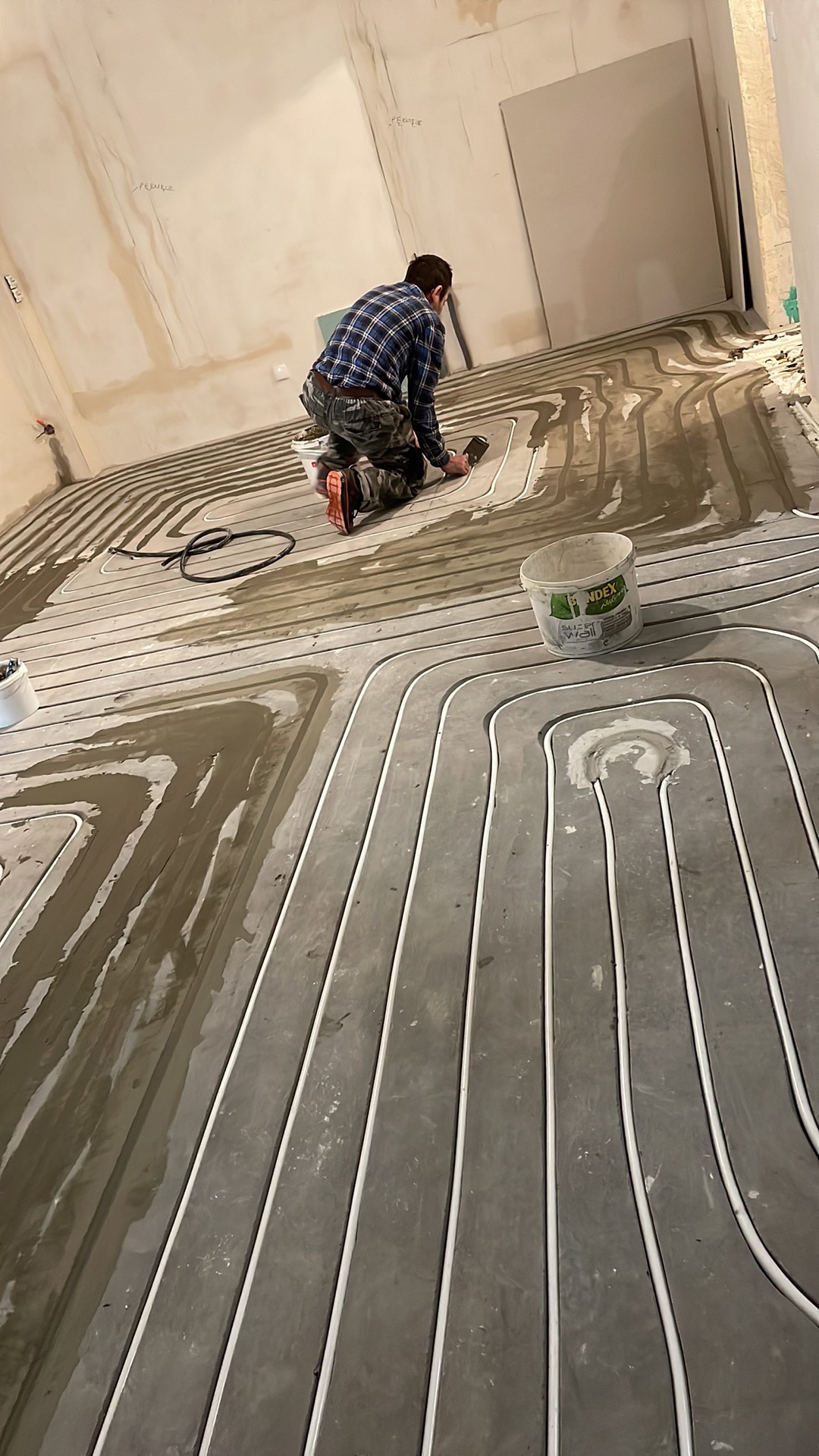 Instalacja ogrzewania podłogowego: mężczyzna w koszuli w kratę rozprowadza wylewkę na rurach w szarej wylewce, widoczny kubeł z napisem 'Index Super Wall'.