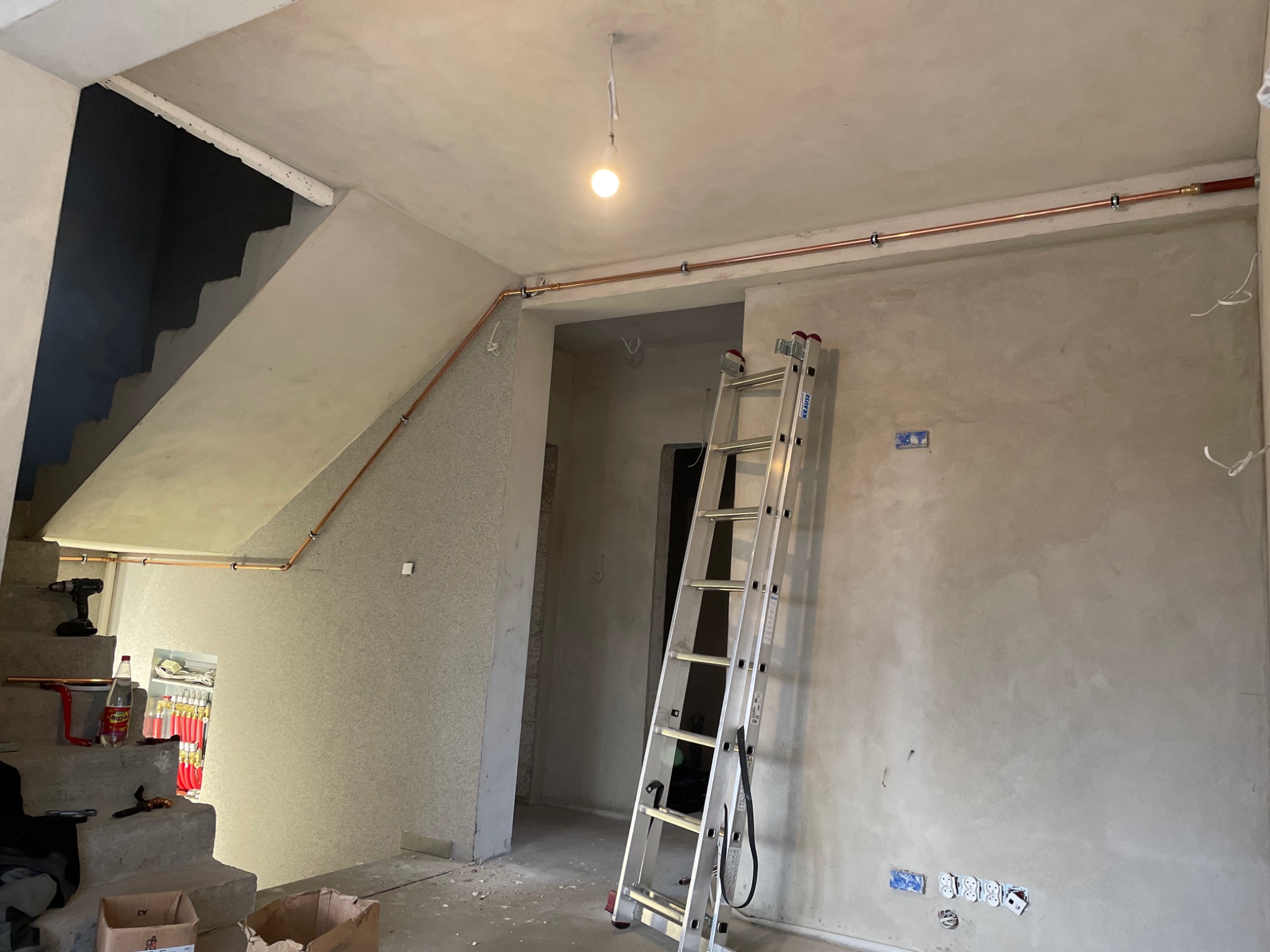 Miedziane rury instalacji grzewczej zamontowane pod sufitem w surowym wnętrzu domu w trakcie budowy, widoczna drabina aluminiowa, schody betonowe i prowizoryczne oświetlenie.