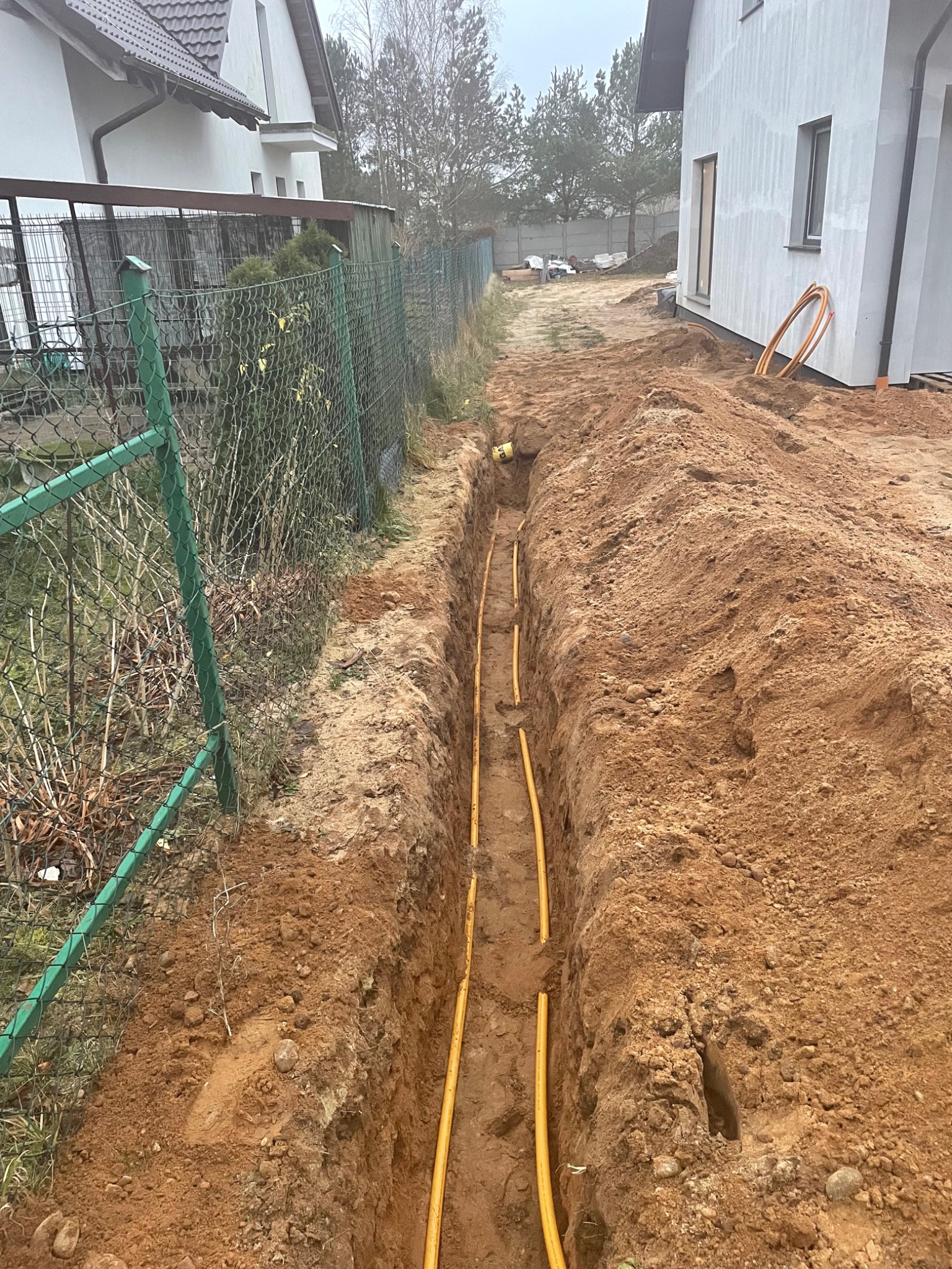Żółte rury gazowe ułożone w wykopie ziemnym wzdłuż ogrodzenia i budynku mieszkalnego w trakcie instalacji.