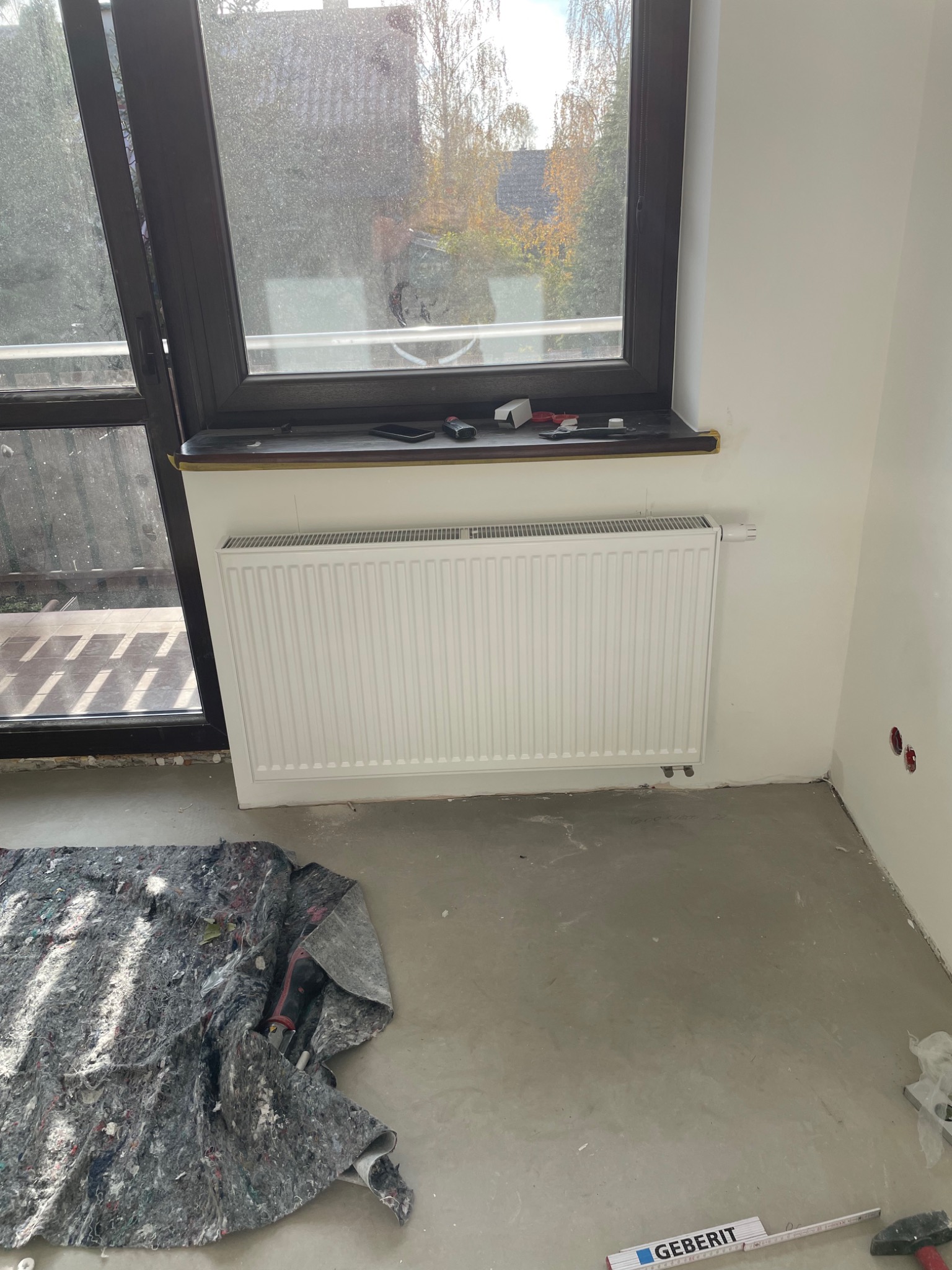 Nowy biały grzejnik panelowy zamontowany pod oknem w surowym wnętrzu, widoczne narzędzia i materiały budowlane na podłodze, ślady prac wykończeniowych.