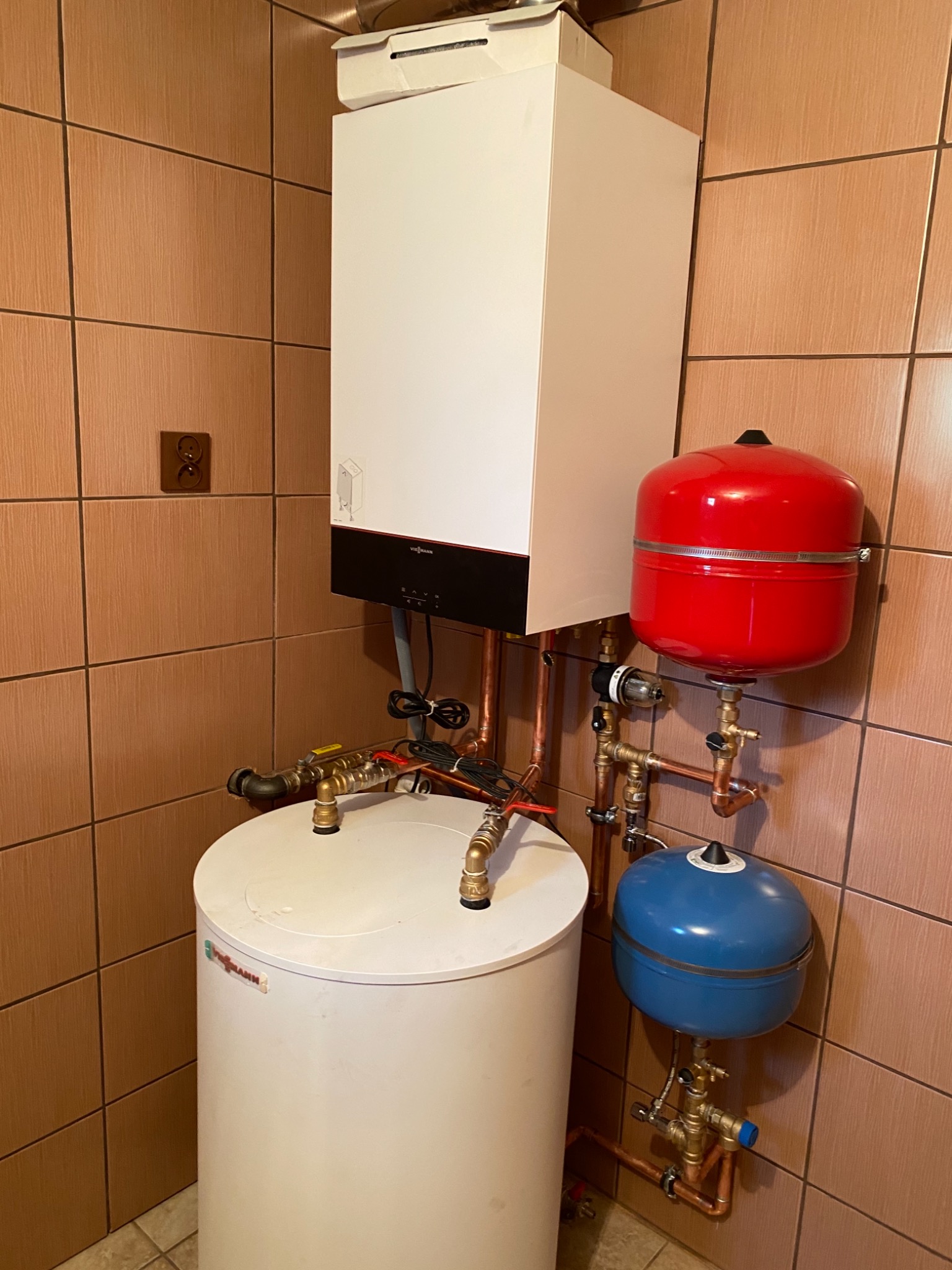 Zainstalowany, wiszący, biały piec gazowy Viessmann z podłączonymi, miedzianymi rurami, czerwonym i niebieskim zbiornikiem wyrównawczym, na tle brązowych płytek ściennych.