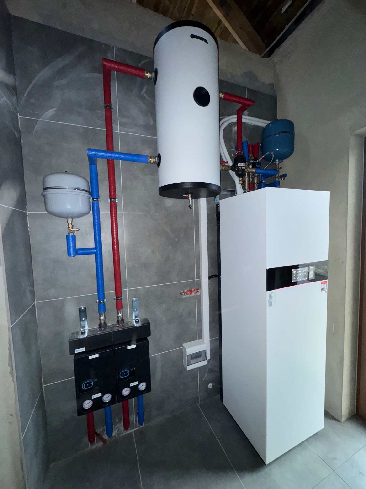 Nowoczesny, biały piec gazowy z podłączonymi rurami w kolorach czerwonym i niebieskim, zainstalowany w pomieszczeniu z szarymi płytkami. Widoczny zbiornik wyrównawczy.