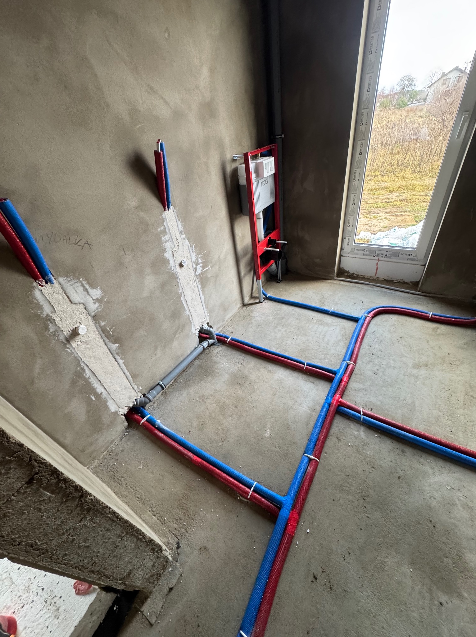 Instalacja wodna z rur PEX w kolorach czerwonym i niebieskim, częściowo zamaskowana w ścianie, z widocznym stelażem podtynkowym WC i oknem w tle.